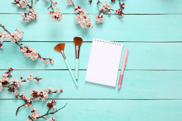 Makeup brushes with notebook and beautiful white flowering branches on blue wooden background. Springtime, beauty concept. Flat lay, top view.