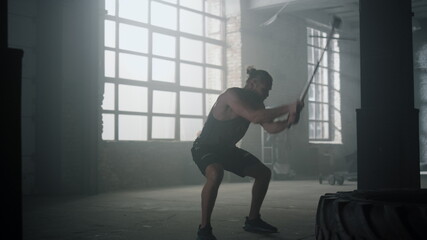 Fototapeta na wymiar Sportsman hitting tire with hammer in gym. Man exercising with sledgehammer
