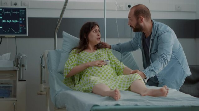 Young woman going into labor in hospital ward sitting with husband holding hand. Caucasian patient doing breathing exercices while having contractions asking for medical assistance