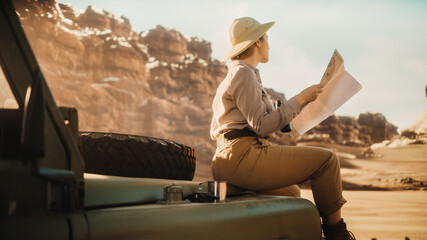 Desert Road Trip: Beautiful Female Traveler Checking Map while Sitting on a Hood of Her Offroad SUV, Planning Route full of Wonder and Exploration. Woman Adventurer Traveling Through Canyon © Gorodenkoff