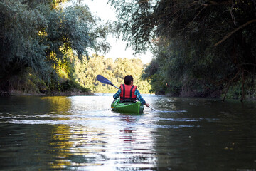Back view on woman in green kayak paddles at wilderness river near trees at summer evening