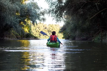 Foto op Canvas Back view on woman in green kayak paddles at wilderness river near trees at summer evening © watcherfox