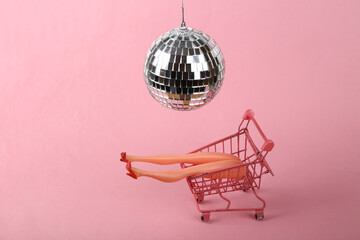 Doll legs in shopping trolley under disco ball on pink background. Minimalism party concept