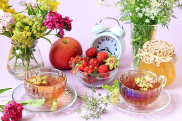 The concept of a good summer morning. Cups of tea among bouquets of wild flowers, strawberries in a cup, jars of jam, apples, alarm clock, light background, close-up, side view