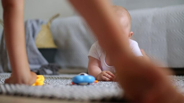Crying infant girl wearing white closing having tummy time on floor near sofa in room, child being upset, faceless kid gives toys to cheer up the baby,