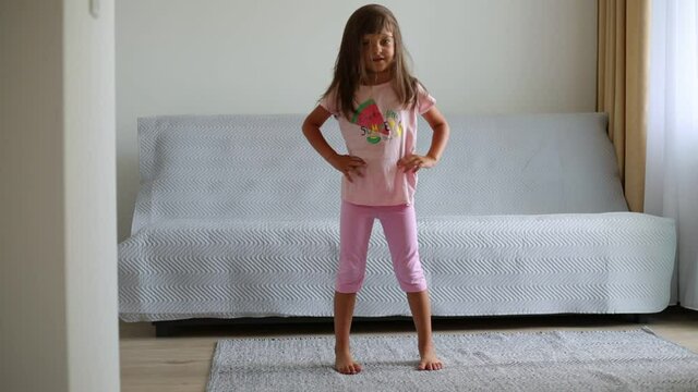 Funny adorable little girl with dark hair wearing pink clothing doing sport alone at home in living room, turning body to sides and smiling, keeping hands on hips.