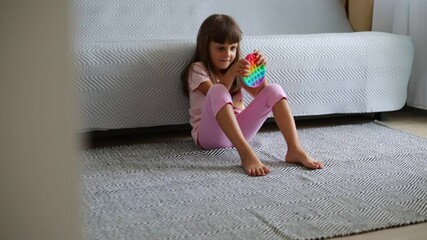 Little cheerful girl with dark hair plays with multi-colored plastic pop it, anti stress toy with bubbles, smiling female kid enjoying modern new popit.