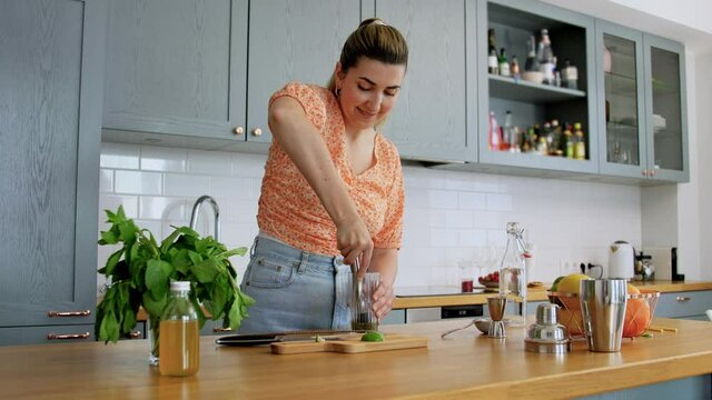 drinks and people concept - happy smiling young woman with pestle making mojito cocktail with lime and peppermint at home kitchen