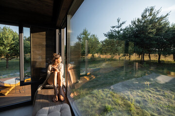 Young woman resting at beautiful country house or hotel, sitting on the window sill enjoying beautiful view on pine forest. Concept of solitude and recreation on nature