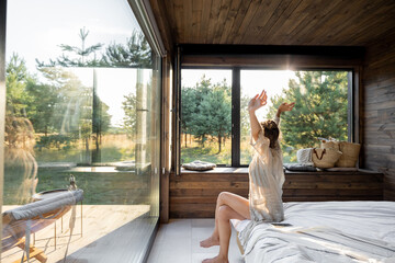 Woman wakes up in a country house or hotel with panoramic windows in pine forest raised her hands yawning. Good morning and recreation on nature concept