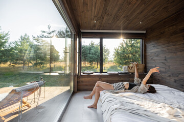 Woman wakes up in a country house or hotel with panoramic windows in pine forest lying on the bed and raised her hands. Good morning and recreation on nature concept
