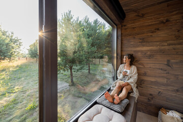 Young woman resting at beautiful country house or hotel, sitting with phone and cup on the window sill enjoying beautiful view on pine forest. Concept of solitude and recreation on nature
