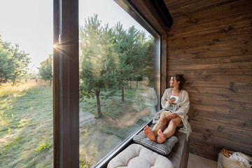 Young woman resting at beautiful country house or hotel, sitting with phone and cup on the window...