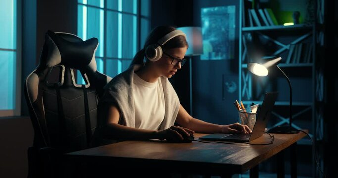 Teenage girl gamer wearing headphones using laptop to play computer games late at night. Schoolgirl spends evening time by doing favorite hobby.