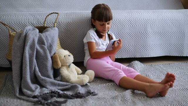 Little girl using video chat on smart phone, preschooler kid sitting on floor near sofa and soft dog toy and talking via video call with friend, expressing positive emotions.