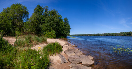 Sandy beach with large cobblestones, stones on the banks of the beautiful wide blue river Narva under the blue summer sky.