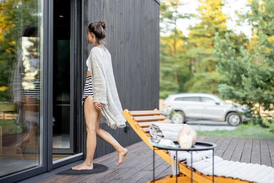Young woman enter the house from the terrace with pine forest view and car on background. Concept of solitude and recreation on nature. Wellness and mindful resort. Beautiful quiet place for vacation.