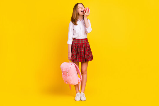 Full body photo of nice little girl eat apple wear shirt skirt bag sneakers isolated on yellow background