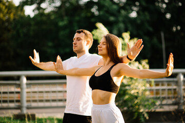 Young couple man and woman meditate together on sunset while standing, yoga on city lawn, summer...