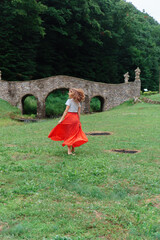 Portrait of a happy Caucasian blonde middle-aged woman with long hair in a white T-shirt and a red sundress with her back to the camera against the background of an old stone bridge. Active leisure.