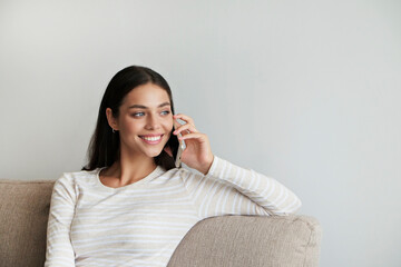 Portrait of young brunette woman wearing white shirt talking on the phone and smiling. Joyful...