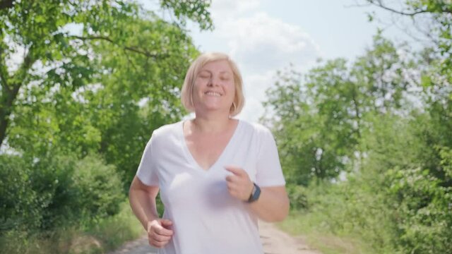 Close up video of the happy woman running along the forest path looking at the video camera with smiling face.