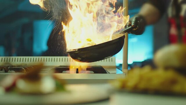 Professional chef cook flambe style . Prepares dish meat in a pan with open flames . Chef frying mixing food on gas hob in commercial kitchen . Man frying steak in flaming pan on hob in restaurant .  
