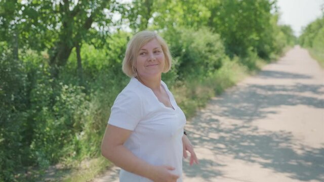 Nice blonde middle-aged woman making fitness running along the path straighten hair and turning towards the camera waving the hand.