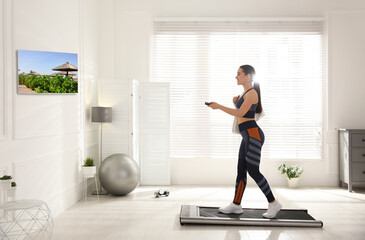 Fototapeta na wymiar Sporty woman with remote control training on walking treadmill and watching TV at home