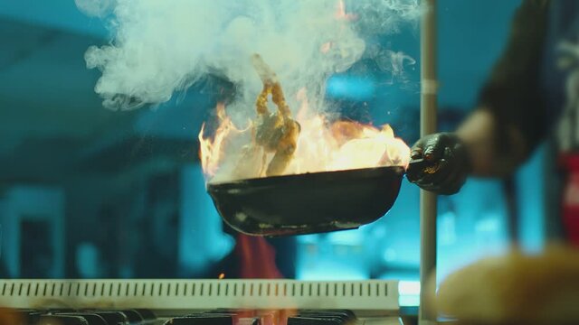 Professional chef cook flambe style . Prepares dish meat in a pan with open flames . Chef frying mixing food on gas hob in commercial kitchen . Man frying steak in flaming pan on hob in restaurant .  