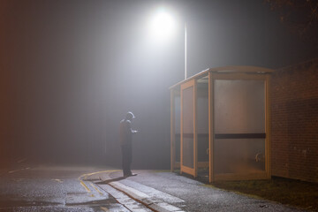 A moody hooded figure looking at a phone next to a bus stop. On a foggy winters evening.