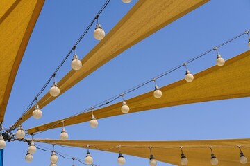 Dynamic airy texture of yellow fabric stretch roof made of canvas sail with rows of bulb lamps for party night lighting on blue sky background.