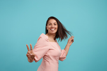 Cheerful smiling european woman with dark hair in pink dress dances, smiles broadly, feels lively and energetic, makes victory gesture, spends free time on disco party, moves against blue background.