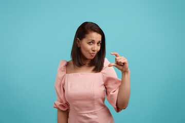 Confused european woman with dark hair in pink dress shows small size with fingers, tells about...