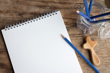 notebook and pencil with small wooden cross with jar of blue pencils on old wooden desk with copy...
