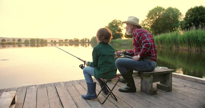 Back view of the old fisherman with teenage boy sitting at the lake and angling while talking early in the morning. Family relationships concept