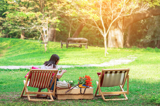 Back View Of Asian Woman Sitting On Garden Chair And By The Table In Garden. Summer Vacation In Green Surroundings. Happy Person Outdoors Relaxing On Deck Chair In Garden. Outdoor Leisure.