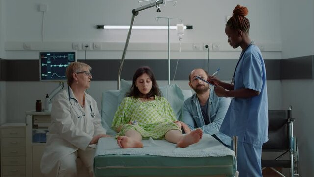 Multi ethnic medical team talking to pregnant woman sitting with husband in hospital ward maternity. Doctor and african american nurse prepare expecting young patient for child delivery