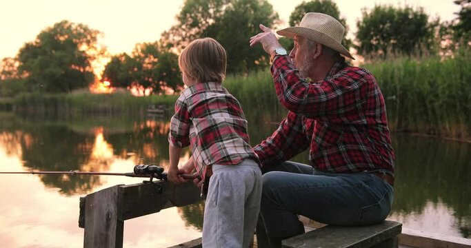 Back view of the grandfather and grandson catching fish at the picturesque nature on a summer day at the sunset. Family relationships and hobbies concept