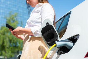 Woman plugging in the charger into her white electric car. Using a smartphone while waiting