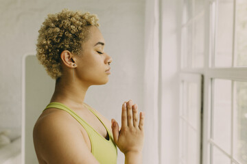 Fototapeta na wymiar Young afro-american female practicing meditation at home. Cute aframerican woman with closed eye standing in padmasana pose. Yoga and meditation concept