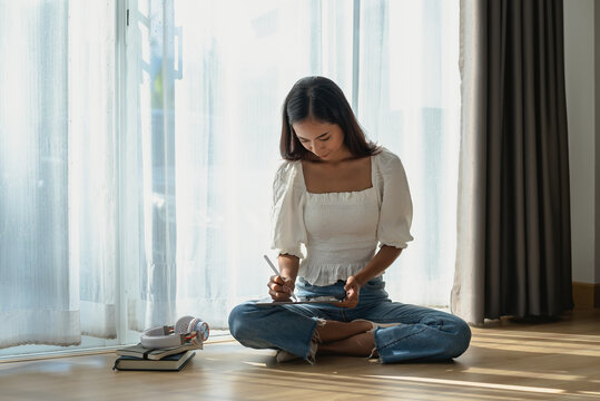 Image of an Asian woman sitting in the living room holding a stylus using a tablet headphones and a book placed in the home .