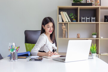 Image of a happy businesswoman with a beautiful smile to work sit in an office chair and taking notes on the laptop the documents are placed at the table.
