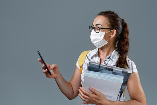 Young woman with brown hair wearing glasses, wears protective surgical mask,has yellow backpack,holds books and uses mobile phone.Portrait of female student in covid times standing on gray background