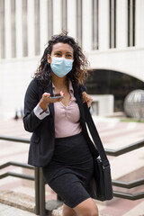 Young hispanic woman with face mask using her smart phone outdoors. Enterprising person. Business during the Covid-19 pandemic.
