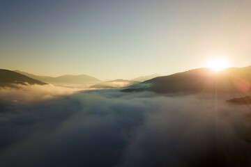Obraz premium Aerial view of vibrant sunrise over white dense fog with distant dark silhouettes of mountain hills on horizon.