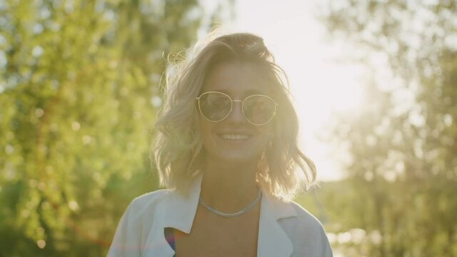 Portrait of young woman in sunglasses standing in forest. Blonde sportswoman smiling, wearing swimsuit, white shirt. Female surf coach looking in camera with smile. Nature, outdoor sports, sunny day