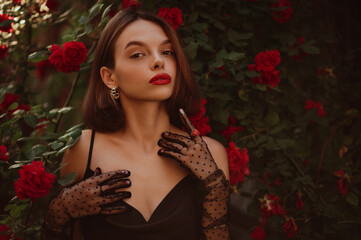 Fototapeta na wymiar Beautiful fashionable woman with red lips makeup wearing trendy polka dot tulle gloves, golden earrings, black dress, posing outdoor, in blooming rose garden. Copy, empty space for text 