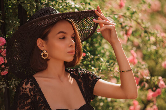Fototapeta Summer street style, fashion portrait of beautiful brunette woman wearing stylish earrings, black wicker hat, lace blouse, posing in street, near blooming roses. Copy, empty space for text 