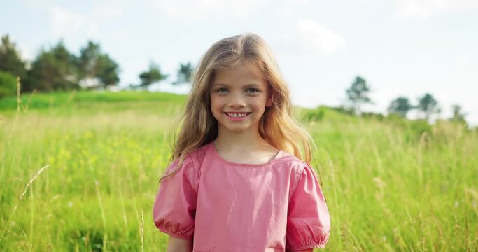Happy girl looking at the camera on green field in summer village. Little child feeling happy on flowering meadow. Female kid spending summer vacation in countryside
